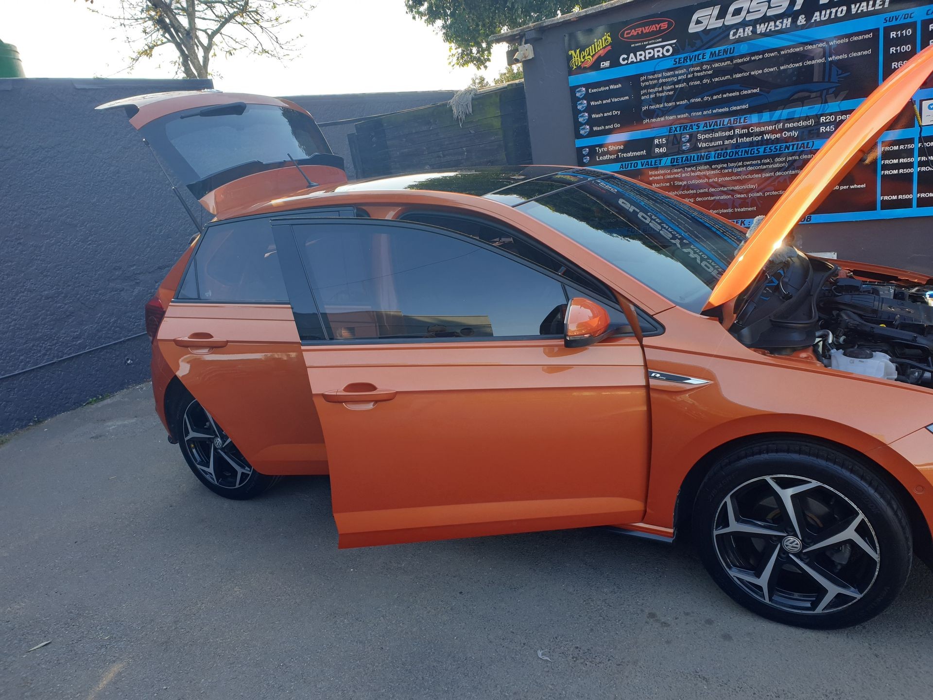 Orange car with open doors and hood near a car wash pricing board.